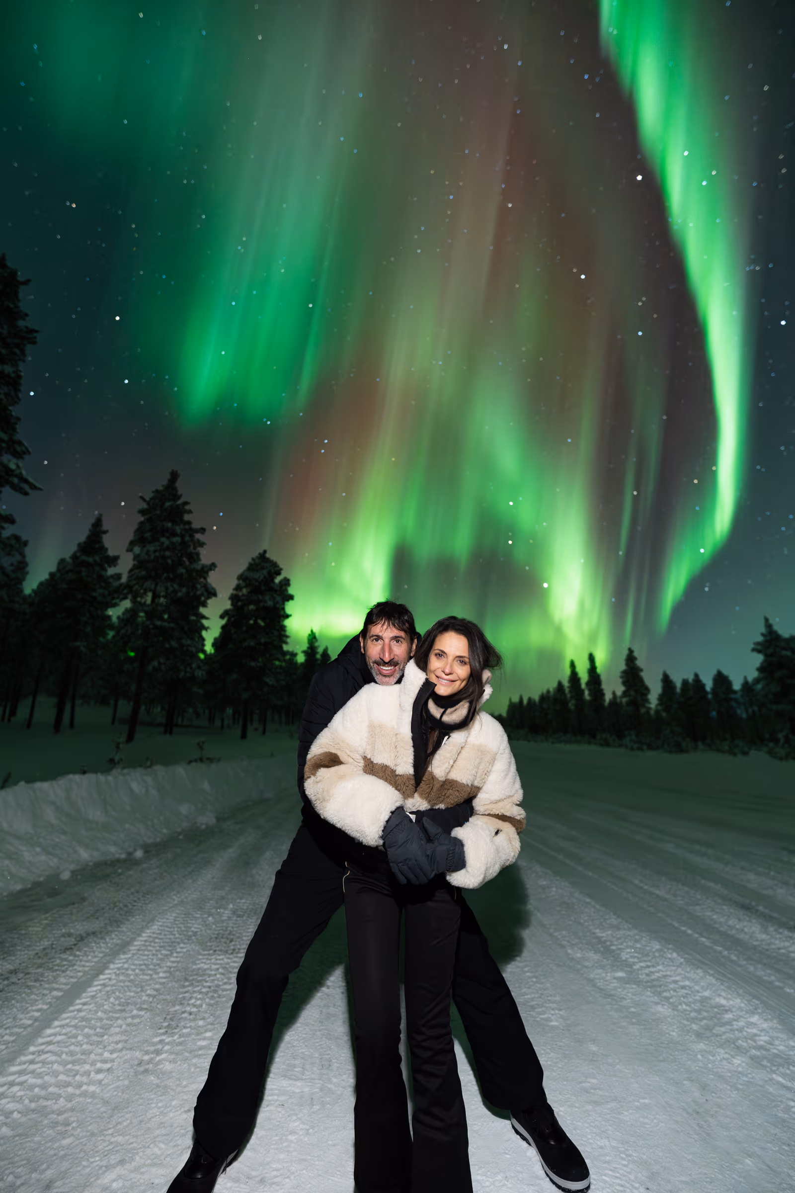 Couple on a snow-covered road beneath green and red northern lights in January Lapland