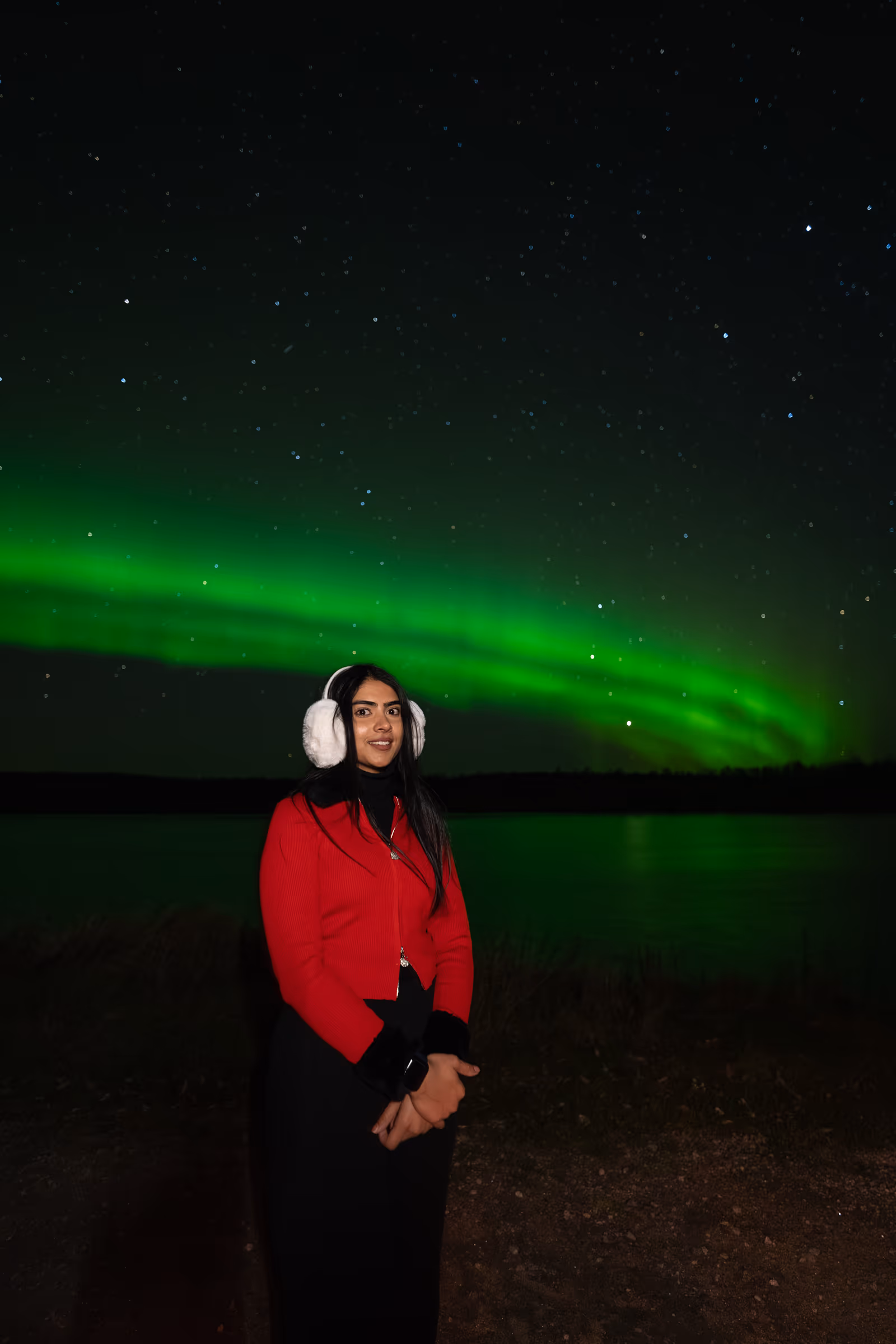 Woman in a red jacket and white earmuffs beside a lake beneath reflected northern lights in autumn Lapland