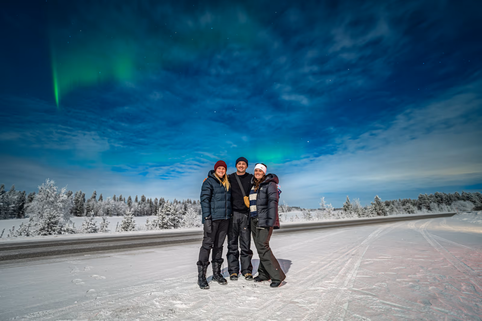 Three friends on a snow-covered road under a bright moonlit blue sky, with a faint aurora overhead